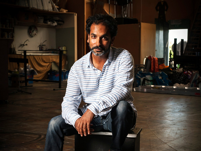 Jacob Rajan sits on a stool wearing a white long shelved shirt with strips and black jeans.