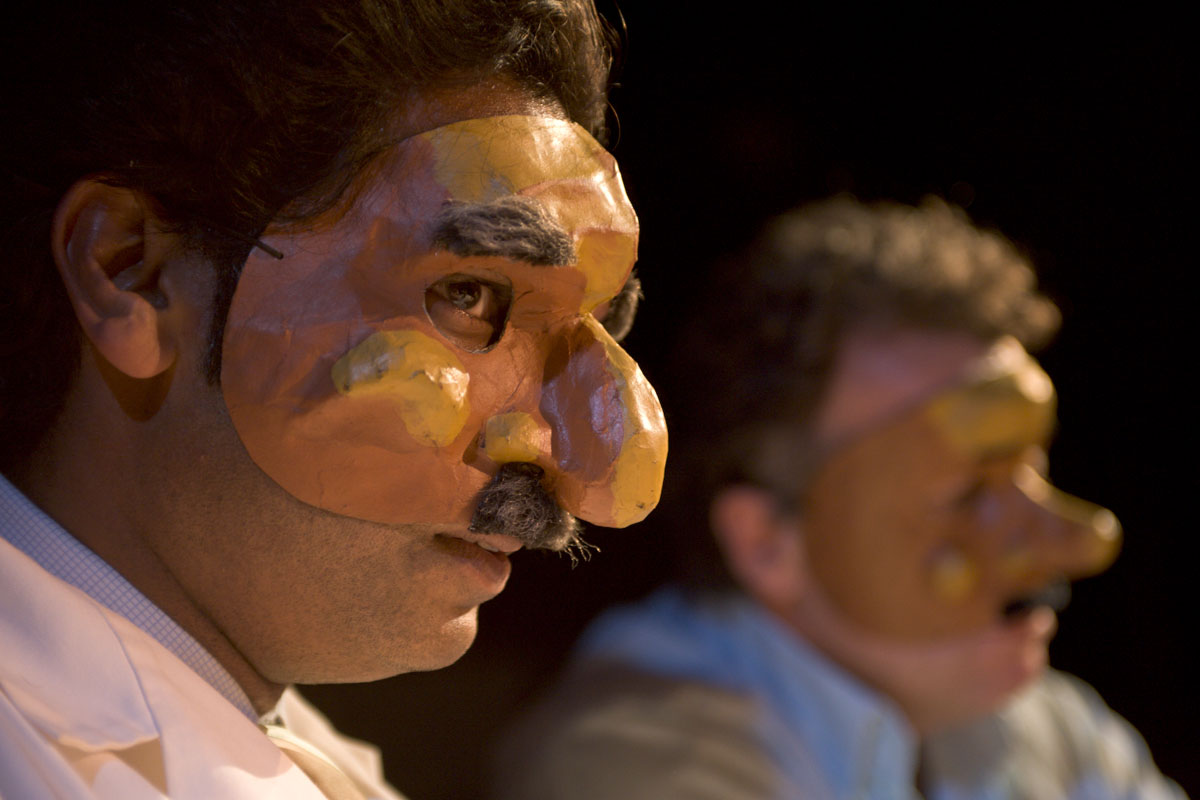 Close up of Jacob Rajan in the foregound wearing a half mask and white clothing. Another actor unfocused in the background wearing a half mask.