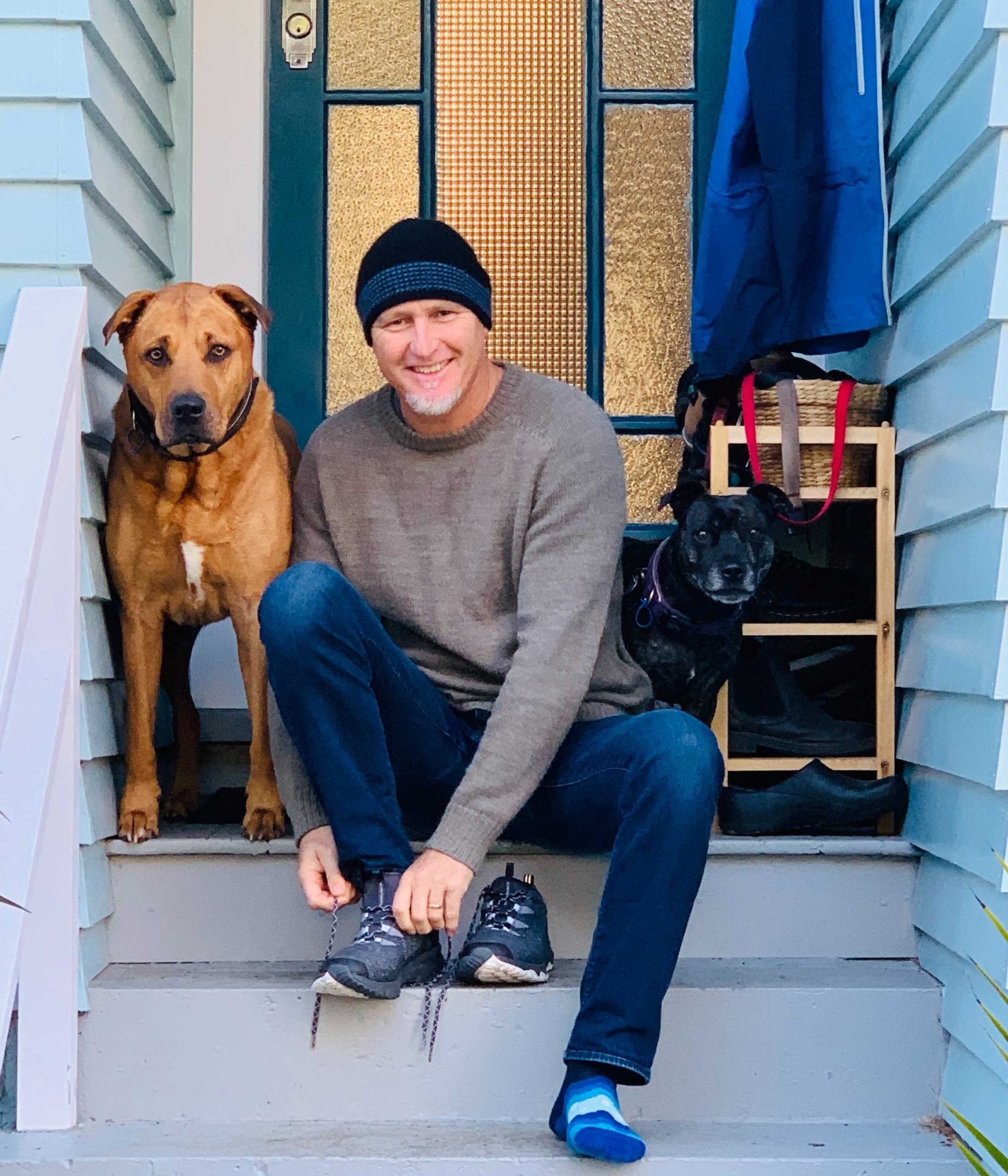 Justin Lewis sits smiling at the camera while tying his sock on his front pouch. HIs tan coloured dog is sitting next to him.