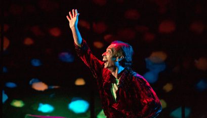 Actor Jacob Rajan reaches for the sky with an expression of joy on his face. The background is black with colorful large light spots that are blue, orange, and red. Jacob is in a velvety red blouse with a green undershirt.