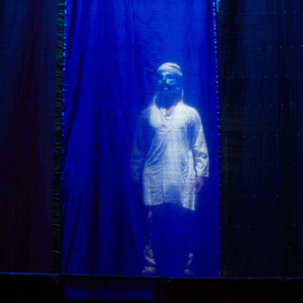 Actor Jacob Rajan in a half mask behind a see through fabric curtain