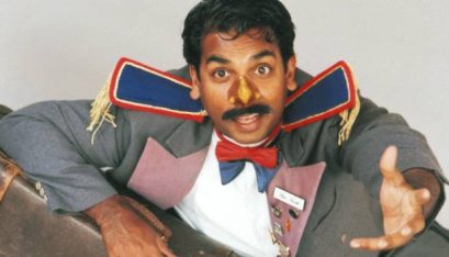 Actor Jacob Rajan wearing a nose mask and dressed as a bell boy holds a suitcase while facing directly up towards the camera on the ceiling