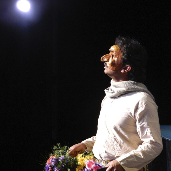 Gobi, played by Jacob Rajan wearing a half-mask, looks up at a bright light representing the moon in the play Krishnan's Dairy. He holds a flowerpot in each hand.