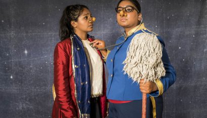 Actor wearing a nose mask and dressed as a bell boy looks concerned as anothe actor dressed as a bell boy holds a mop and listens to the heart of the first actor