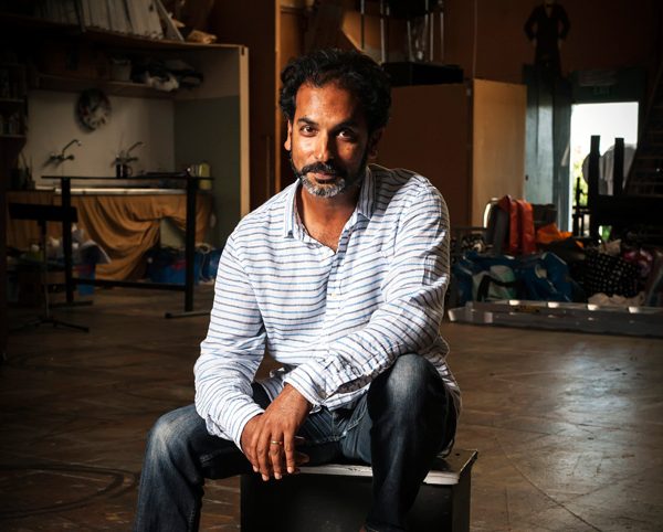 Jacob Rajan sits on a stool wearing a white long shelved shirt with strips and black jeans.