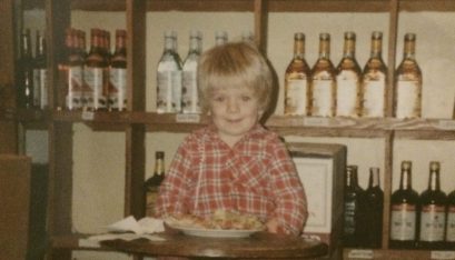 Andrew Potvin as a young toddler in front of a wall of alcohol. Andrew sits on a bar stool with chips on the table in front of him