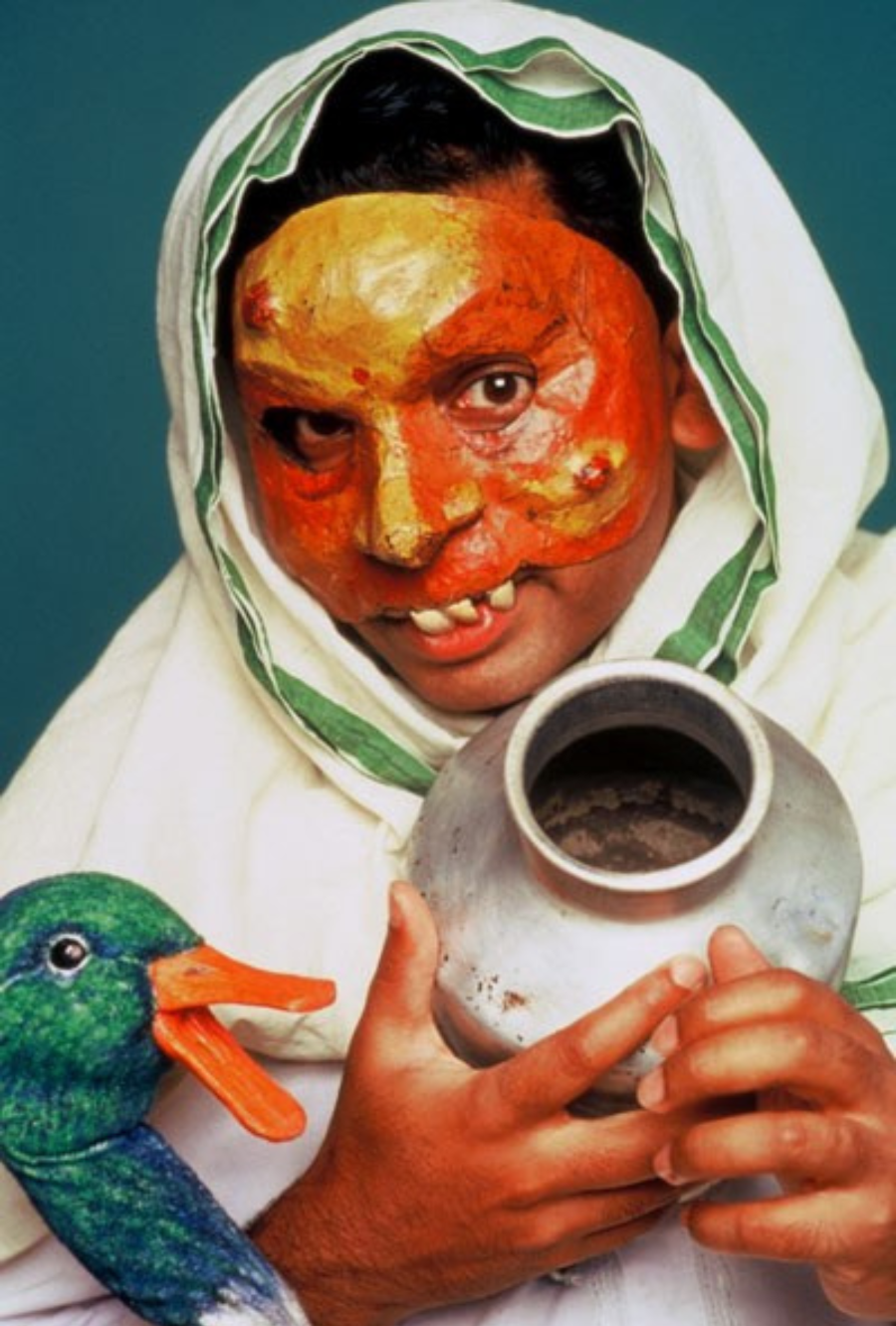Jacob Rajan wears a papier mache half mask with a white shawl draped around him and his head, playing the character Kalyani. He holds a vase and points it at the camera with a shy smile. There is a duck puppet beside Kalyani.