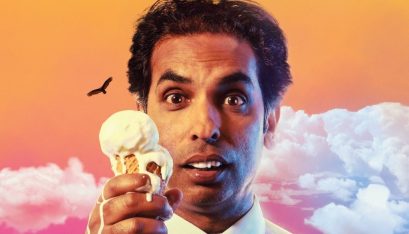 Actor Jacob Rajan looking confused while holding a melting ice cream cone. He is wearing a white collared shirt. The background is a dreamy orange and pink with a silhouette of a bird and lush pink and white clouds.