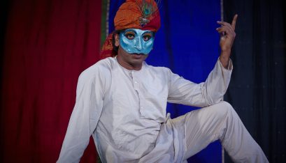 Actor Jacob Rajan sits as Emperor Shah Jahan wearing a blue half face mask in front of colourful background