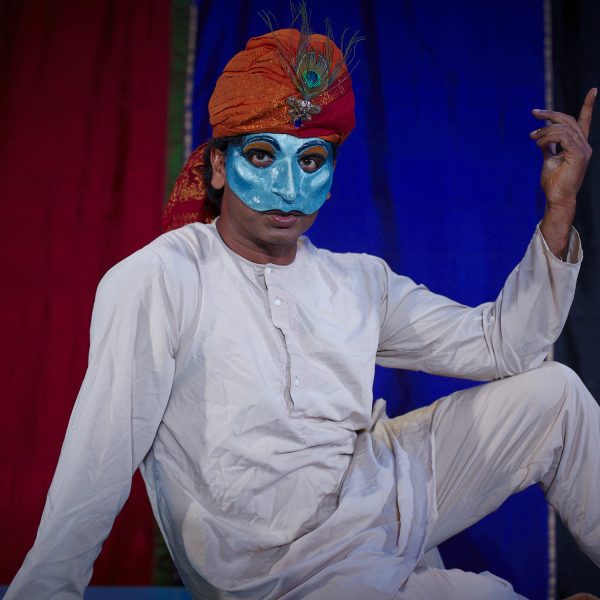 Actor Jacob Rajan sits as Emperor Shah Jahan wearing a blue half face mask in front of colourful background