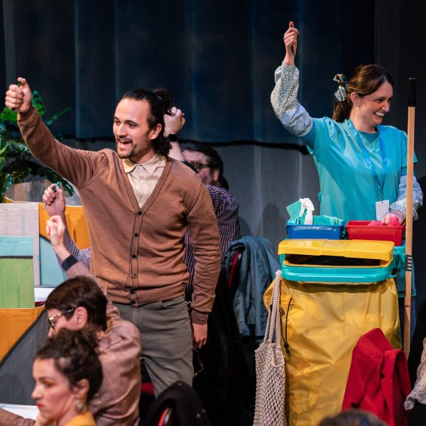 Justin Rogers and Catherine Yates portray Neil and Joy in the comedic play, DIRTY WORK. In a colourful office setting, the characters - Neil in a brown buttoned-up cardigan and Joy in a blue cleaner's uniform - hold their pens high above their head, grinning.