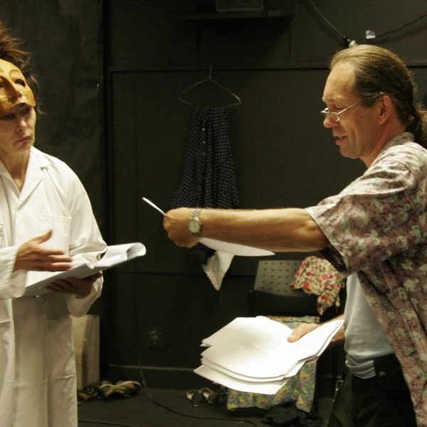 Murray Edmonds stands with script notes that he delivers to an actor wearing a half mask and lab coat.