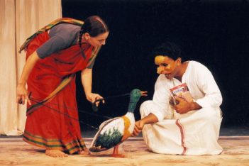 Actor Kate Parker stands leaning over while holding a duck puppet. Actor Jacob Rajan kneels while patting the duck and holding a book.