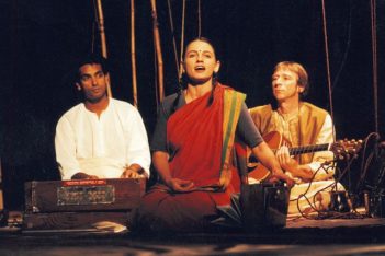 Actor sits kneeling in the foreground while singing. Jacob Rajan sits in the background playing an instrument with another actor next to him playing a gutair.