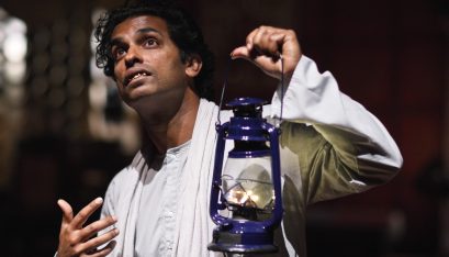 Actor Jacob Rajan stands in front of a white door holding a lantern