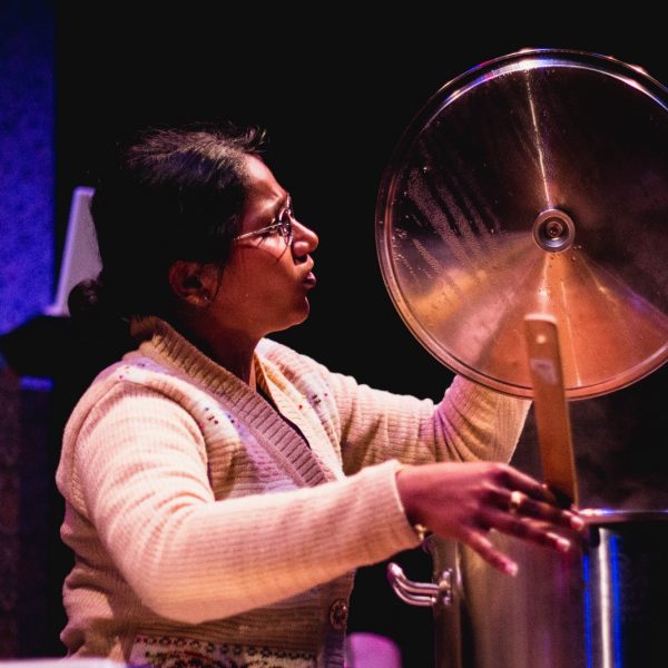 Actor Kalyani as Mrs Krishnan looks into a pot lit which has a light shining on it while she stirs the dahl in the pot