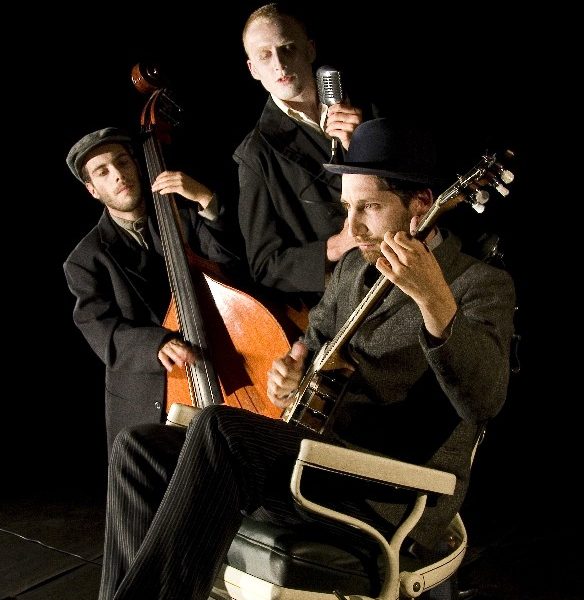 David Ward sits on a chair with a banjo while two other musicians stand with him.