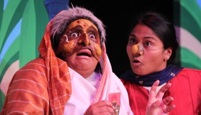 Actor dressed an old lady stands to the left with a half mask one.The other actor is looking shocked at the photo