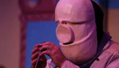 An Actor in an oversized face mask with a circular nose looks intently at their hands