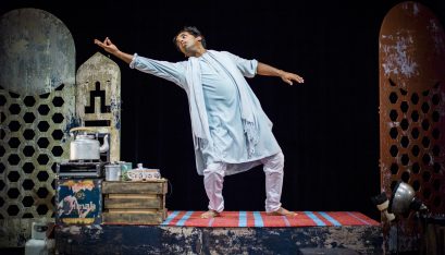 Actor Jacob Rajan stands in a dance pose on a raised set with a tea trolley next to him