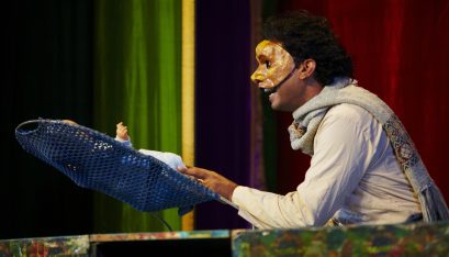 Actor Jacob Rajan as Zina with a half face mask looking over a toy baby in a cradle