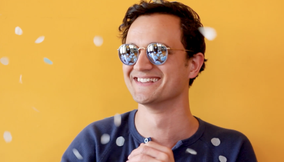 Actor Justin Rogers smiles past the camera in a blue shirt and sunglasses. Behind him is a yellow wall.