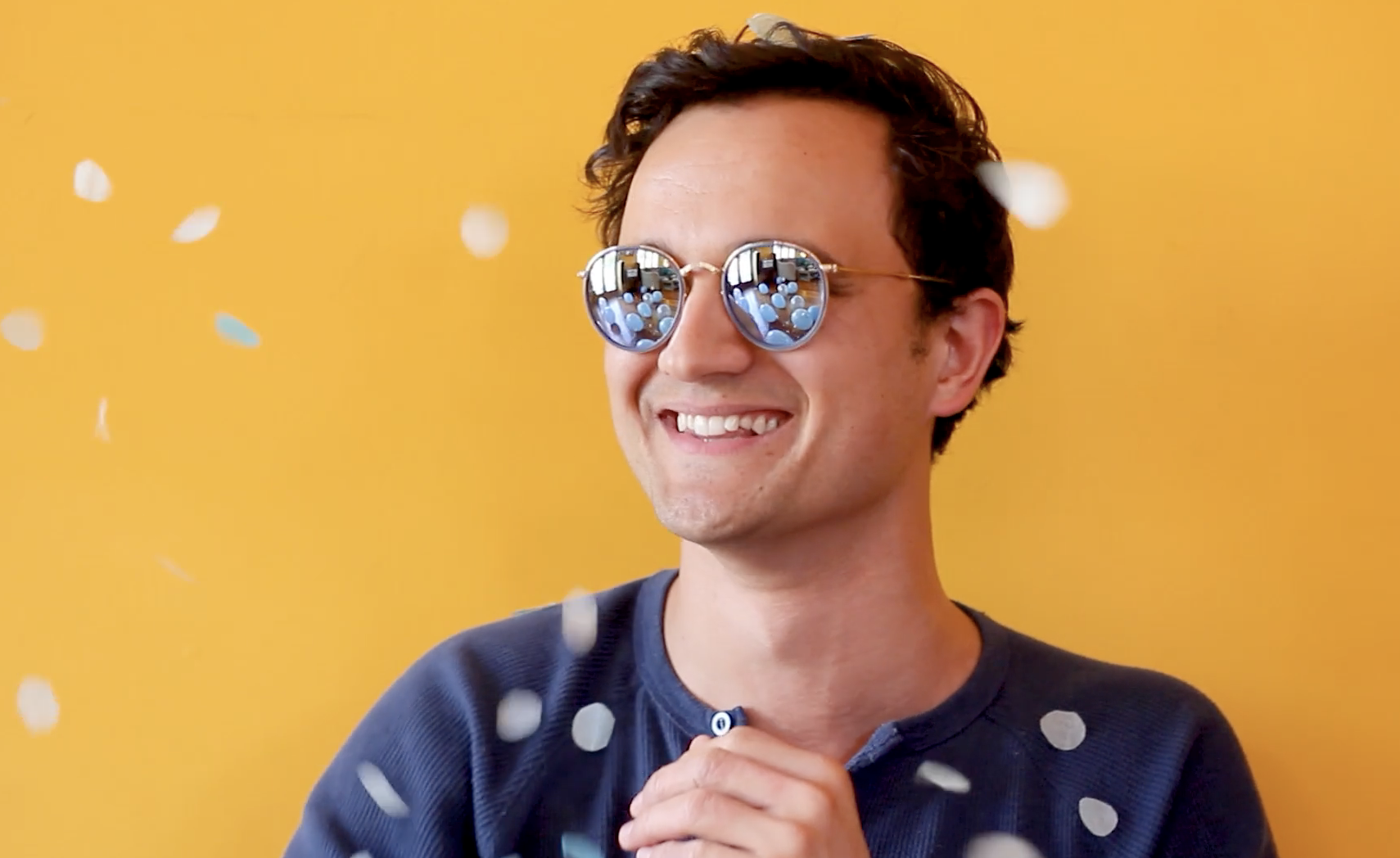 Actor Justin Rogers smiles past the camera in a blue shirt and sunglasses. Behind him is a yellow wall. 