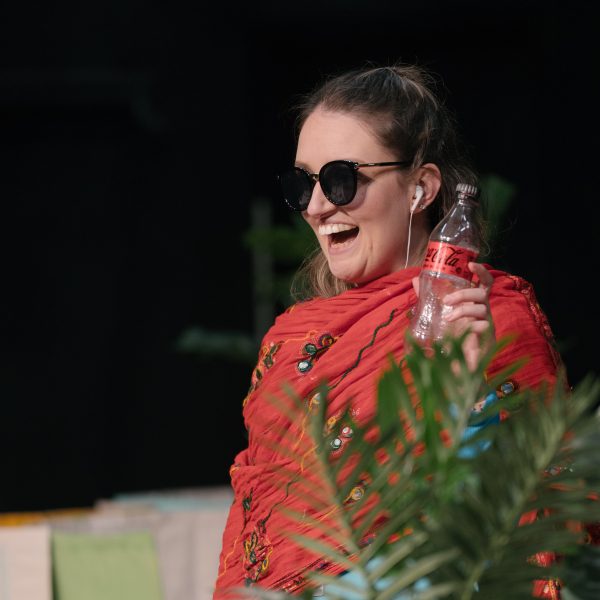 Actor Catherine Yates stands wearing sunglasses and a scarf while holding a coke bottle and grinning towards the audience