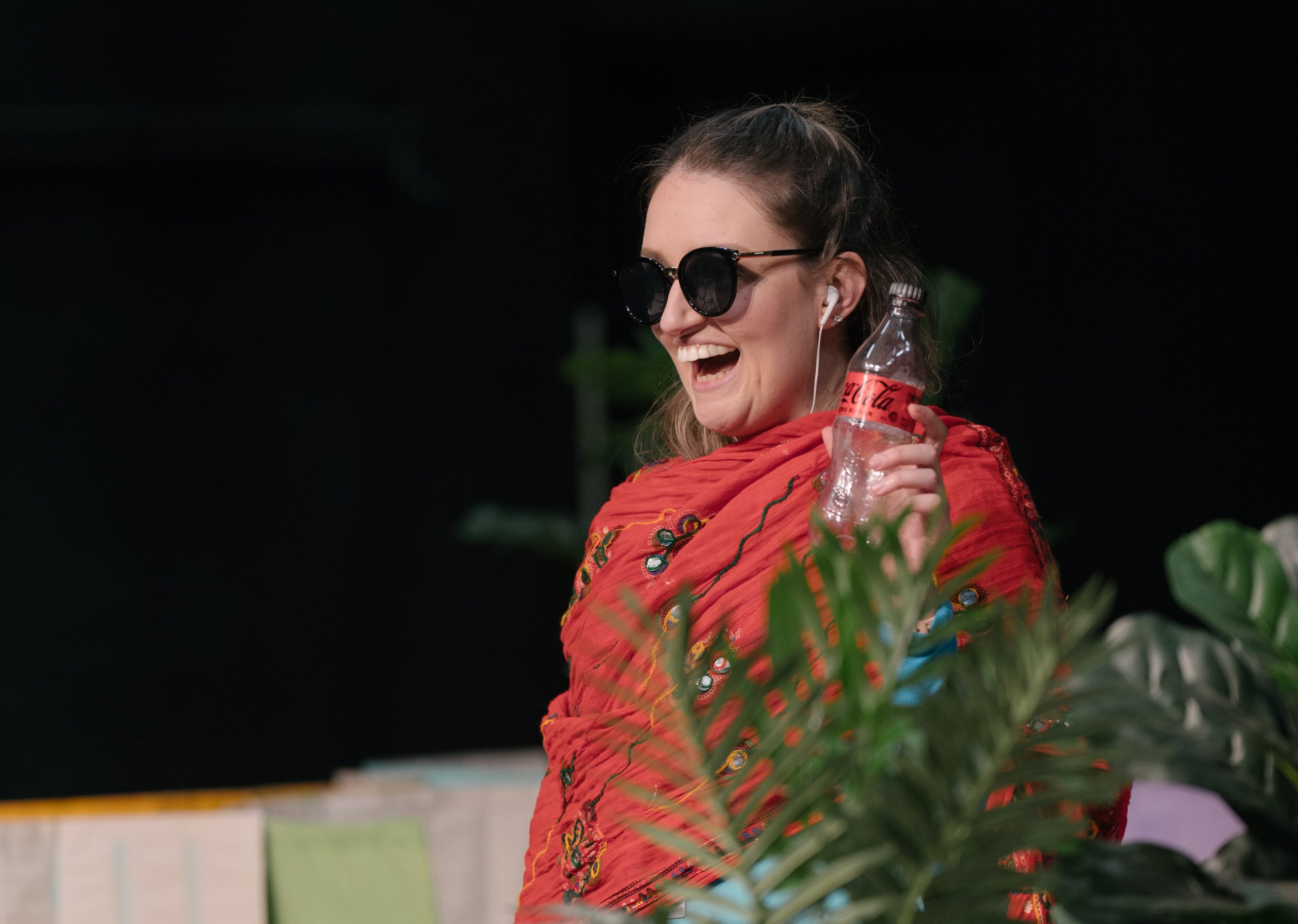 Actor Catherine Yates stands wearing sunglasses and a scarf while holding a coke bottle and grinning towards the audience