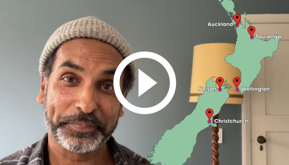 Jacob Rajan sits with a smile on his face and a cartoon map of NZ next to him. The map has dots for Auckland, Nelson, Christchurch, Wellington & Tauranga