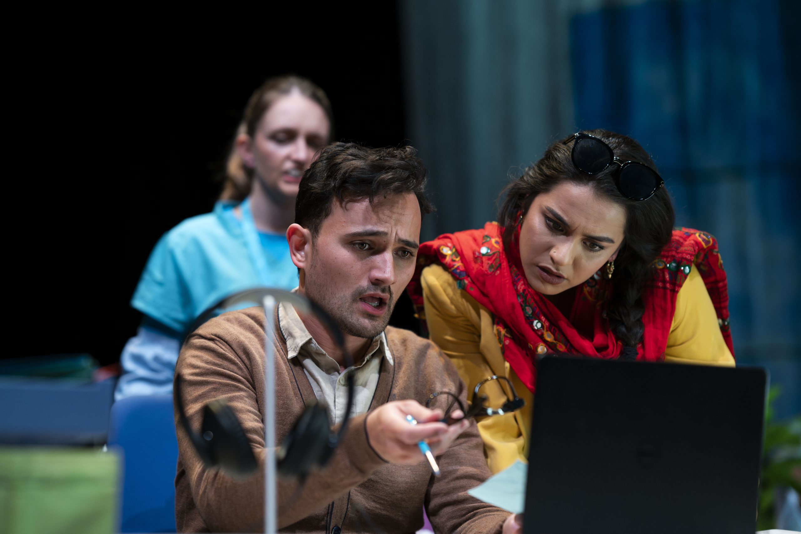 Actor Justin Rogers sits looking confused at his laptop while holding a pen out. Tessa Rao stands looking at a piece of paper in Justin's hand. Catherine Yates is seen blurred in the background