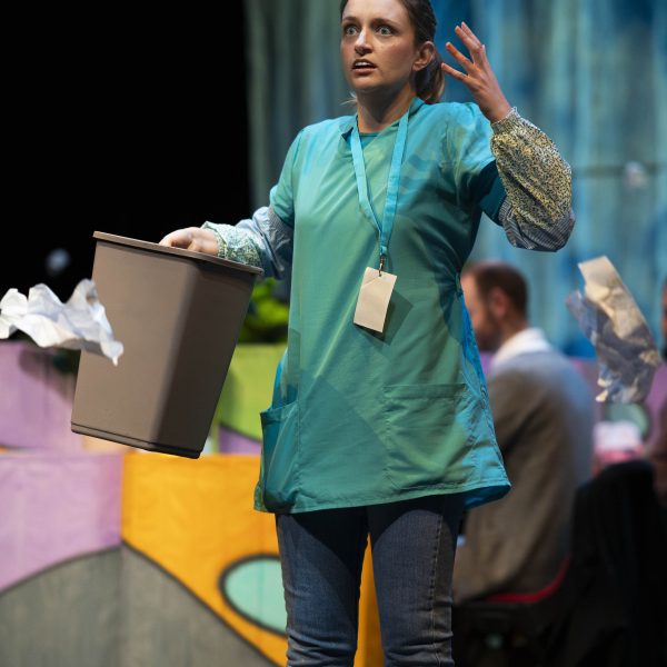 Catherine Yates is dressed in a cleaners outfit standing confused as balls of paper fall around her