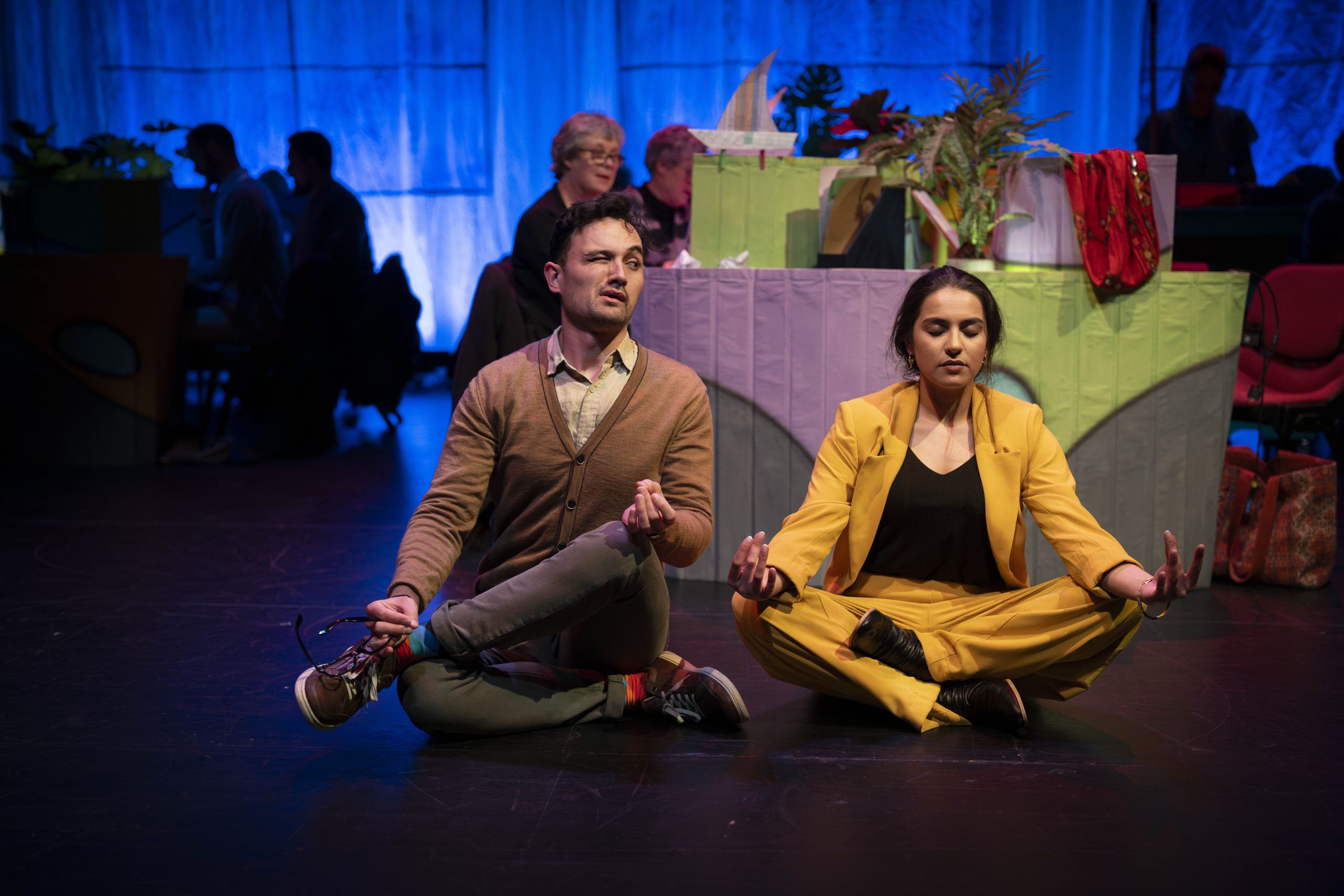 Actor Tessa Rao sits in a yellow pant suit in a yogi pose on the ground. Justin Rogers sits in a brown jumper in an awkward yogi pose while staring at Tessa with one eye open and one closed. 