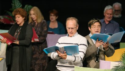 Choir Members stand singing while holding music folders of sheet music.