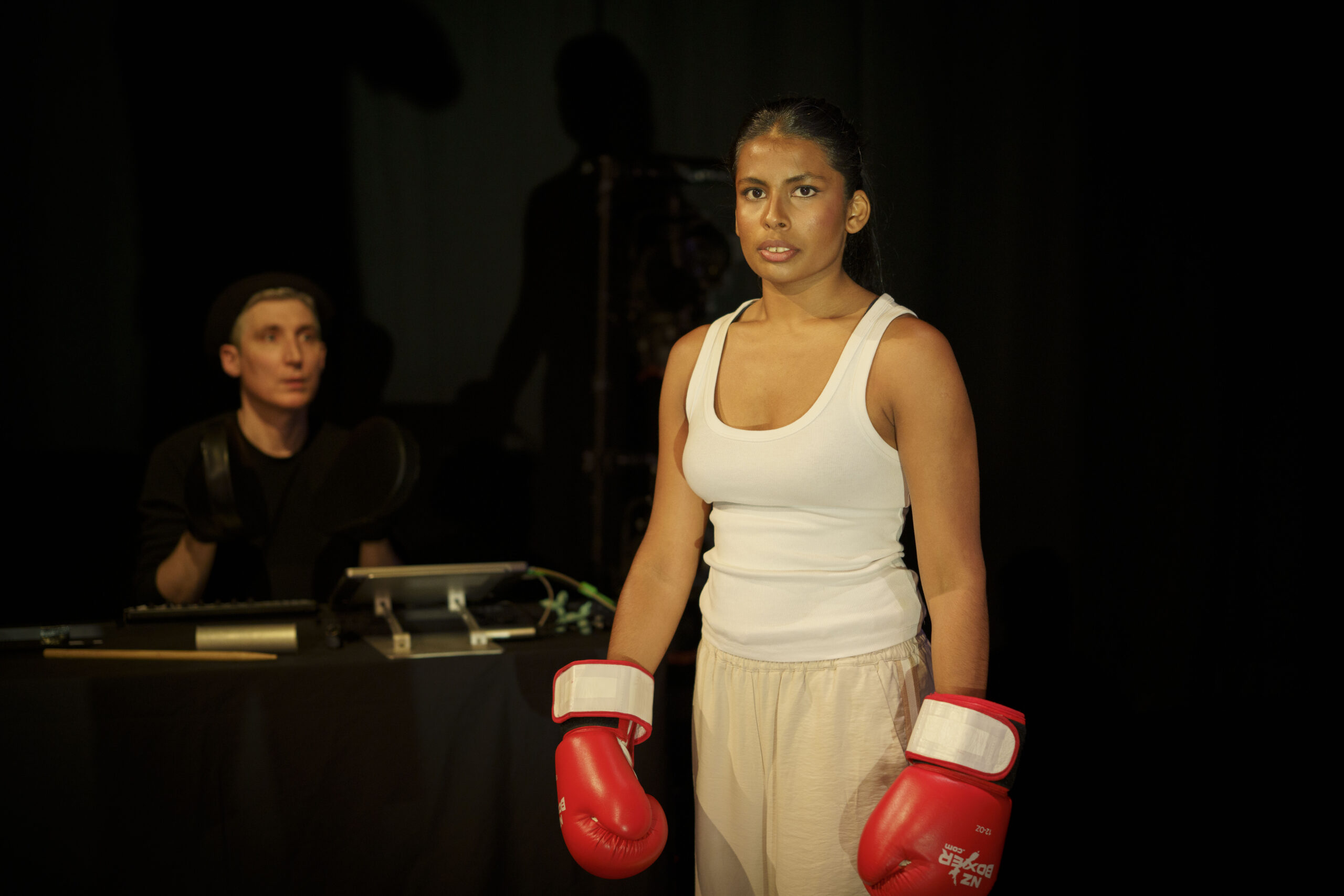 Alisha Jacob stands in workout costume with red boxing gloves. The Musician Dave can be seen in the background
