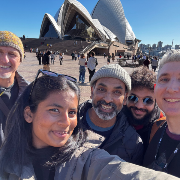 Five smiling adults take a selfie in front of the Sydney Opera House on a sunny day.