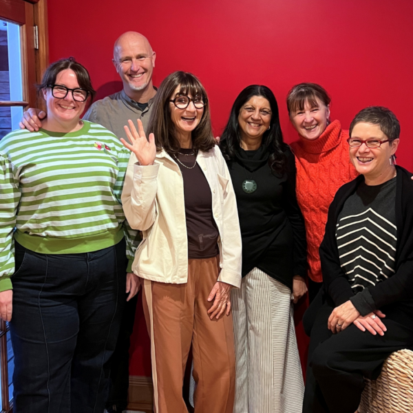 Six smiling adults pose indoors against a red wall; one woman waves at the camera, all in casual clothing.