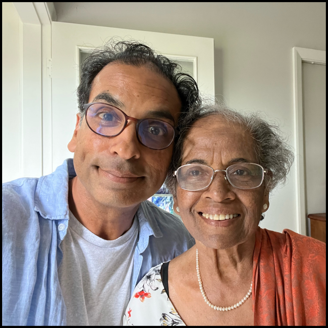 Man and elderly woman wearing glasses, smiling together; she wears a pearl necklace and an orange shawl.