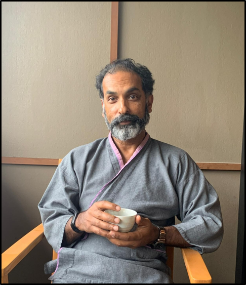 Bearded man in gray robe sitting in wooden chair, holding a small white teacup against a neutral wall.
