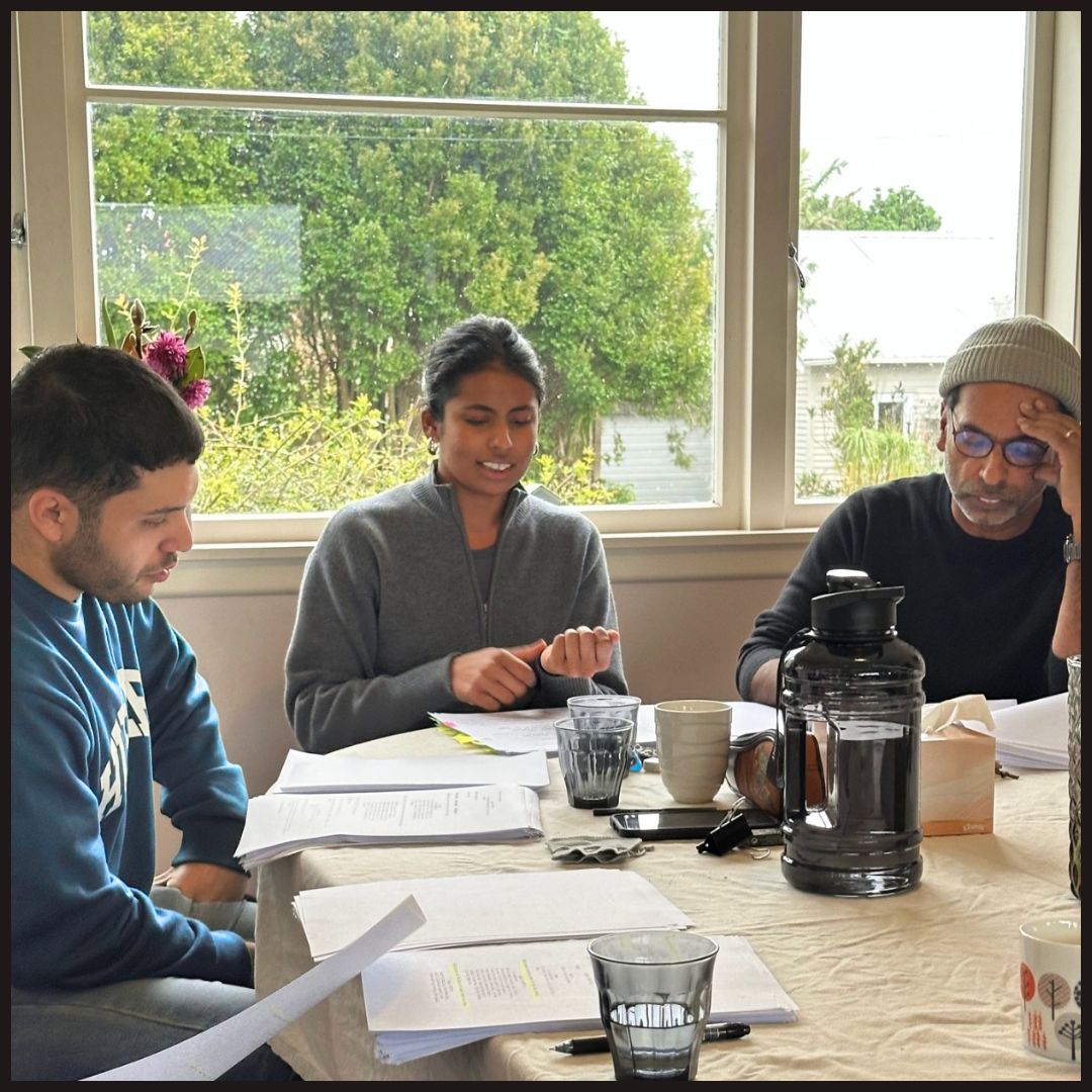 Three people around a table reviewing documents; woman speaking as two men read papers near a window.