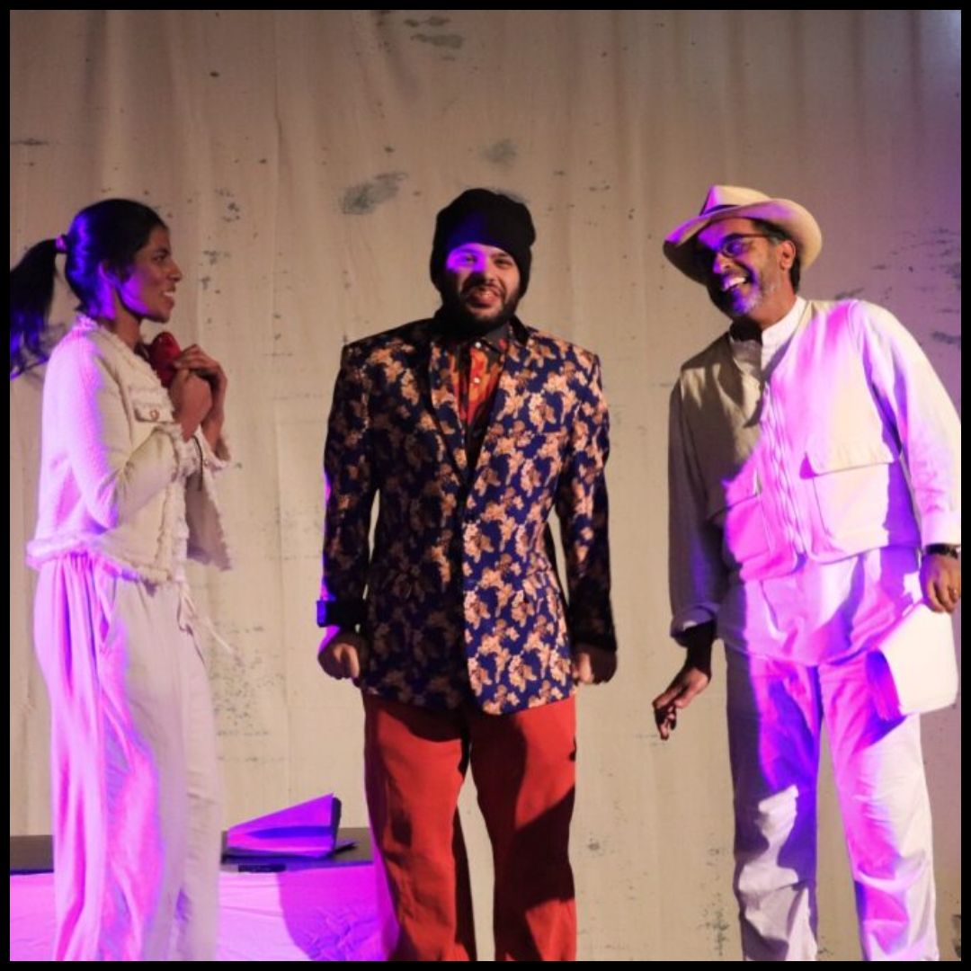 Three actors under colored lights—woman in white with a red prop, center in patterned jacket and beanie, man in white hat.