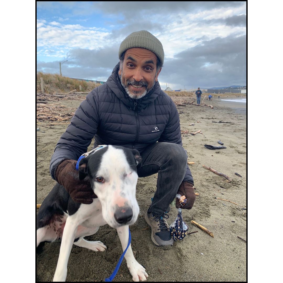 Smiling man in knit hat and puffer jacket crouches on a windy beach holding a black-and-white dog on a blue leash.