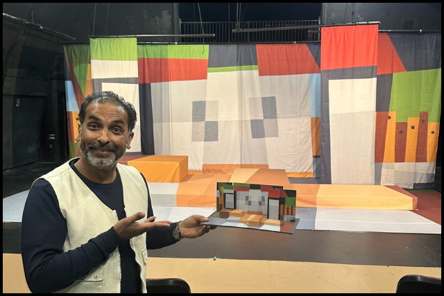 Jake gestures to a small colorful stage model with a matching geometric backdrop and platforms.