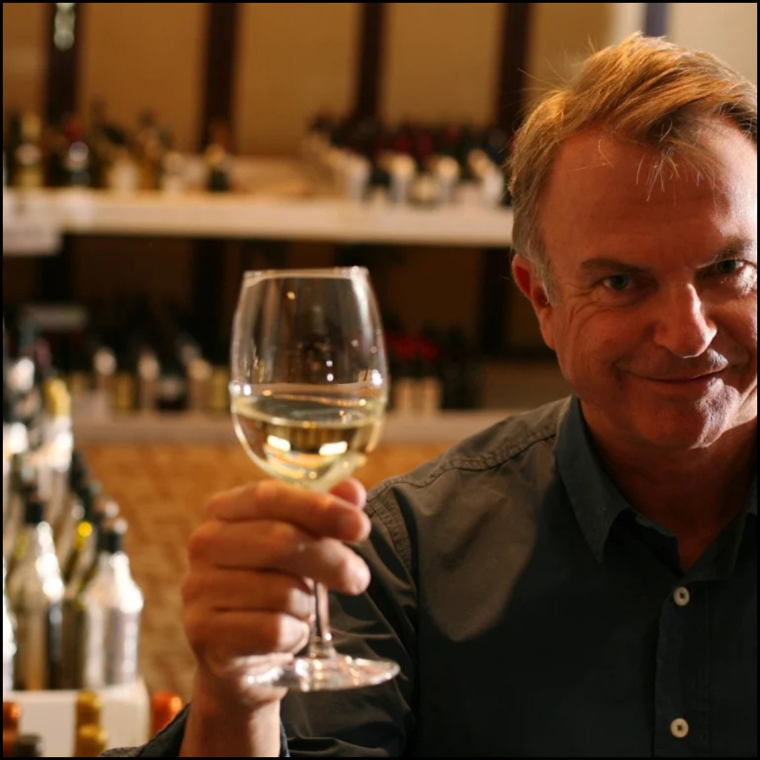 Sam Neil holding up a glass of white wine in a wine shop or tasting room.