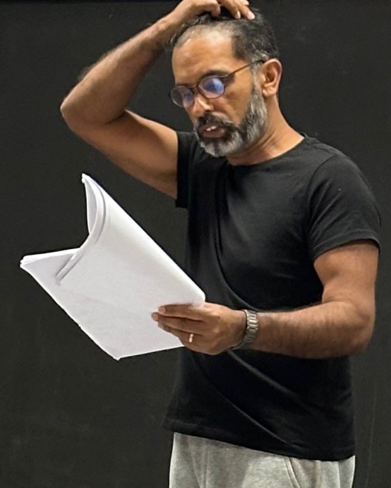 Middle-aged man with glasses reading a stack of papers, holding them with one hand while scratching his head.