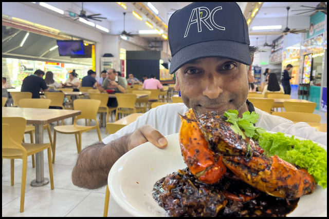 Jake in a black cap holds a plate of glossy black-pepper crab garnished with lettuce in a busy food court.