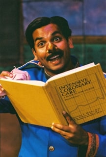 Smiling mustachioed man holding an open yellow medical book, animatedly gesturing as he reads.