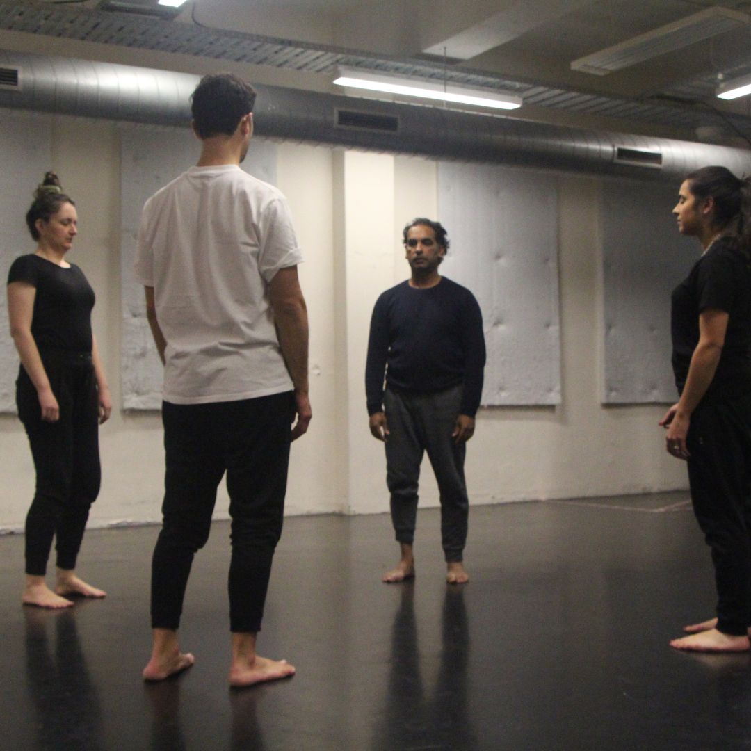 Four people stand barefoot in a studio, forming a loose circle and facing inward.