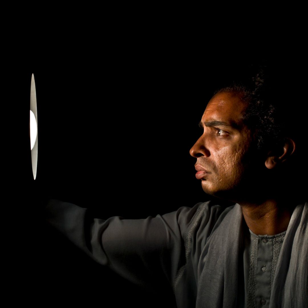 Man in profile lit by a narrow vertical light, reaching toward a small bright opening against a black background.