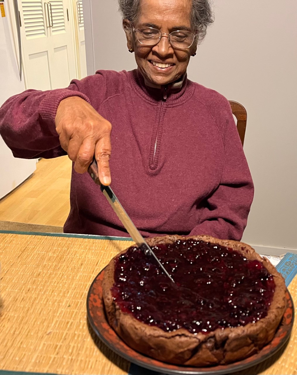 Smiling older woman in maroon sweater slicing a round cake topped with glossy berry jam.